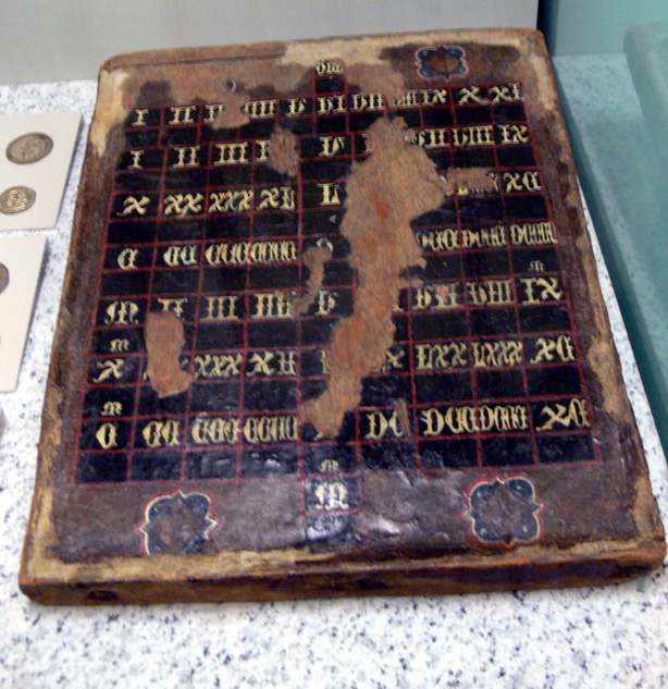 Une planche de bois quadrill&eacute;e de petites cases sombres, chacune marqu&eacute;e d&rsquo;un chiffre romain dor&eacute; ; la peinture est us&eacute;e par endroits, jusqu&rsquo;au bois nu.