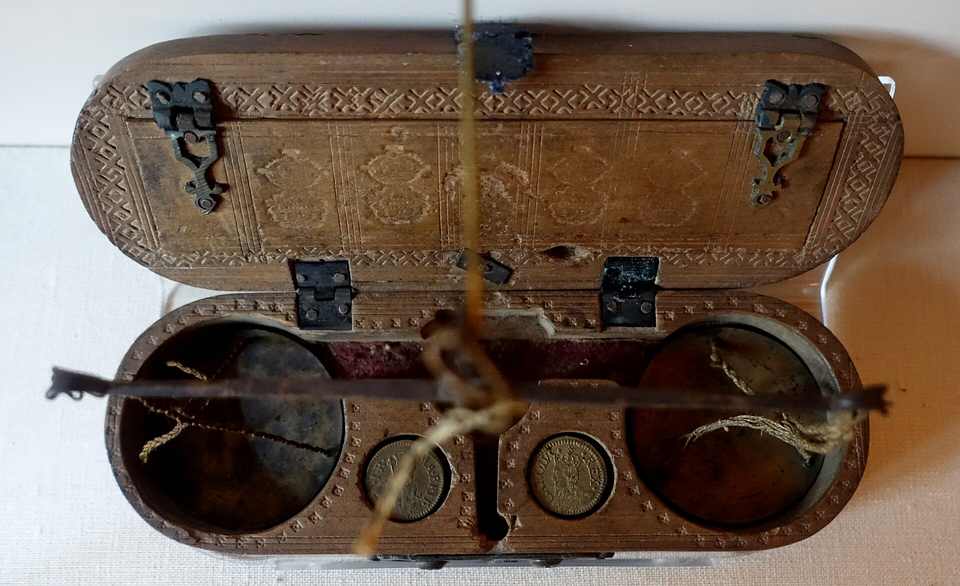 Un &eacute;tui ovale en bois et cuir, orn&eacute; de motifs en losange, ouvert pour montrer une petite balance de laiton suspendue entre deux logements ronds o&ugrave; reposent deux pi&egrave;ces.