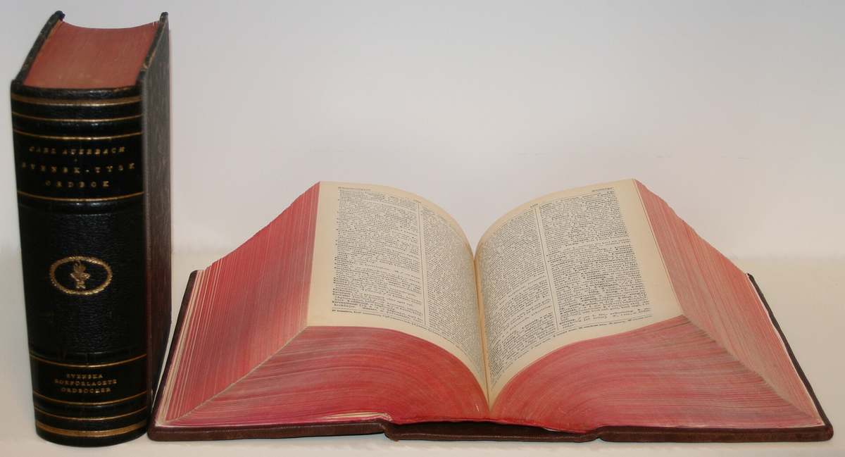 Deux volumes reli&eacute;s de cuir rouge et noir : l&rsquo;un ferm&eacute;, debout ; l&rsquo;autre ouvert &agrave; plat, montrant deux pages denses de d&eacute;finitions imprim&eacute;es sur plusieurs colonnes.