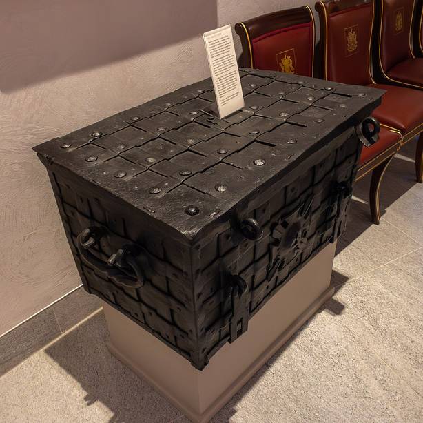 Un coffre en fer noir, ferm&eacute;, pos&eacute; sur un socle : ses parois sont couvertes de plaques rivet&eacute;es et de bandes crois&eacute;es, avec un anneau de chaque c&ocirc;t&eacute; et une petite serrure au centre du couvercle.