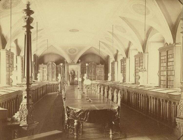 Photographie ancienne en teintes s&eacute;pia d&rsquo;une grande salle de biblioth&egrave;que vout&eacute;e : de hautes &eacute;tag&egrave;res de livres courent le long des deux murs, une longue table centrale est born&eacute;e de chaises, et une silhouette se tient debout au fond, pr&egrave;s d&rsquo;une porte.